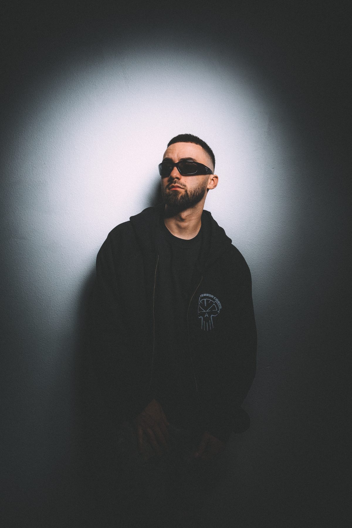 Vlado in sunglasses and a black zip hoodie, leaning against a spotlit wall